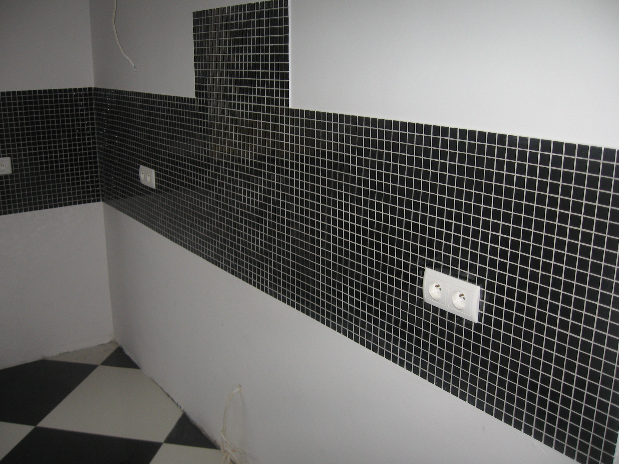 Fragment ściany wyłożonej czarną mozaiką z białymi fugami, z widocznym gniazdkiem elektrycznym; podłoga w czarno-białą szachownicę.